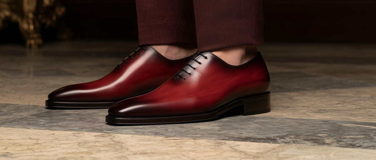 Burgundy hand-painted wholecut oxford shoes with patina, worn with burgundy trousers on a stone floor